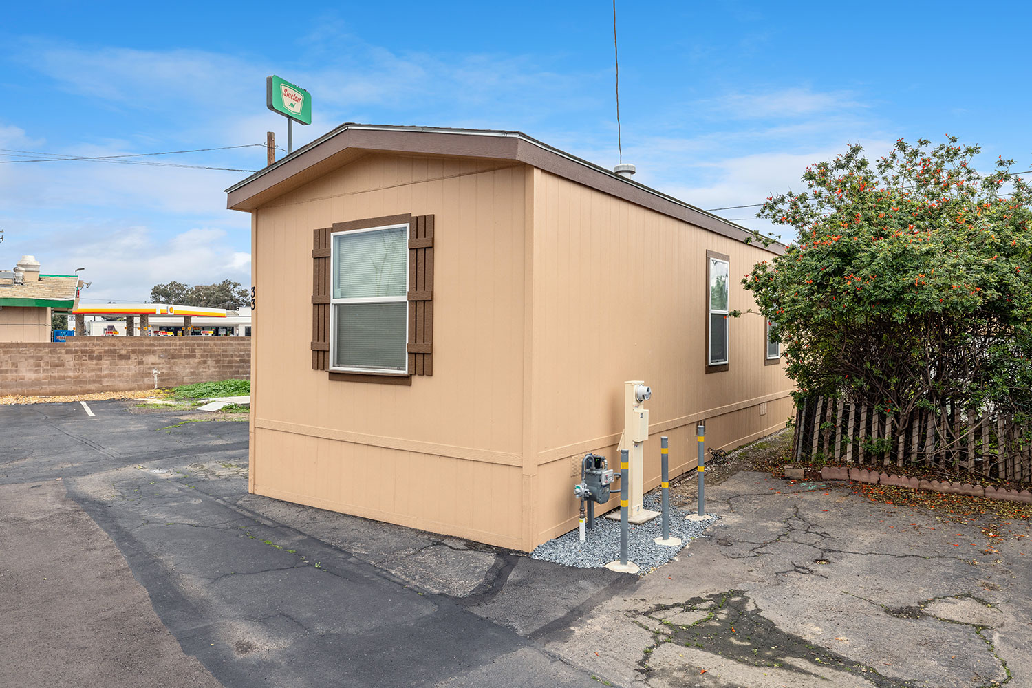 Beige mobile home with brown shutters at Park View Mobile Home & RV Park in Fresno, CA, next to a paved driveway and a flowering bush, with a gas station visible in the background.